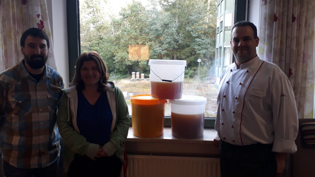 Drei Eimer mit Honig, Voll, dreiviertel voll und halb voll, als Pyramide gestapellt, links davon Dr. Klemens und Frau Dressler, rechts davon Herr Gehring.