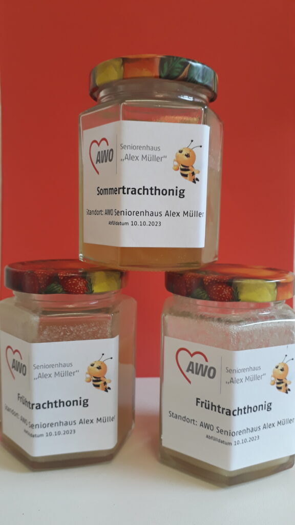 Marmeladengläser zut Befüllung mit Honig, darauf kleine Comic-Biene, AWO Logo "Alex Müller" und Beschriftung Früh- bzw Sommertracht