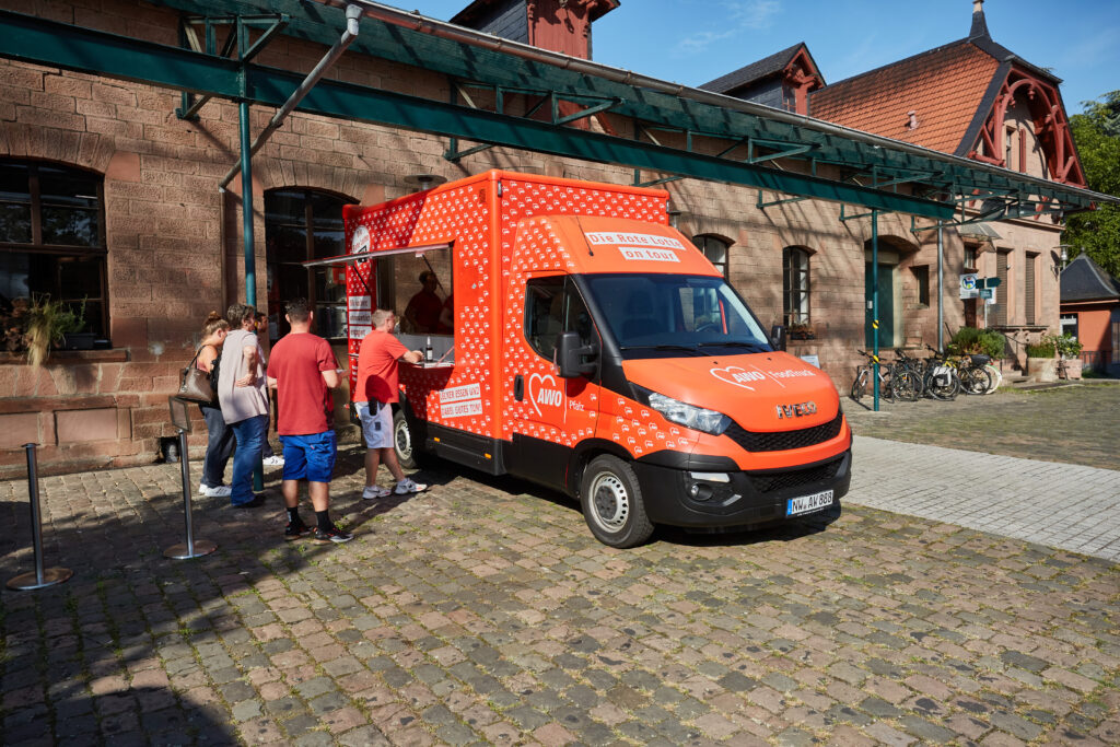 Kleiner roter Foodtruck voller AWO-Herzen. Menschen stehen vor der geöffneten Klappe und warten.